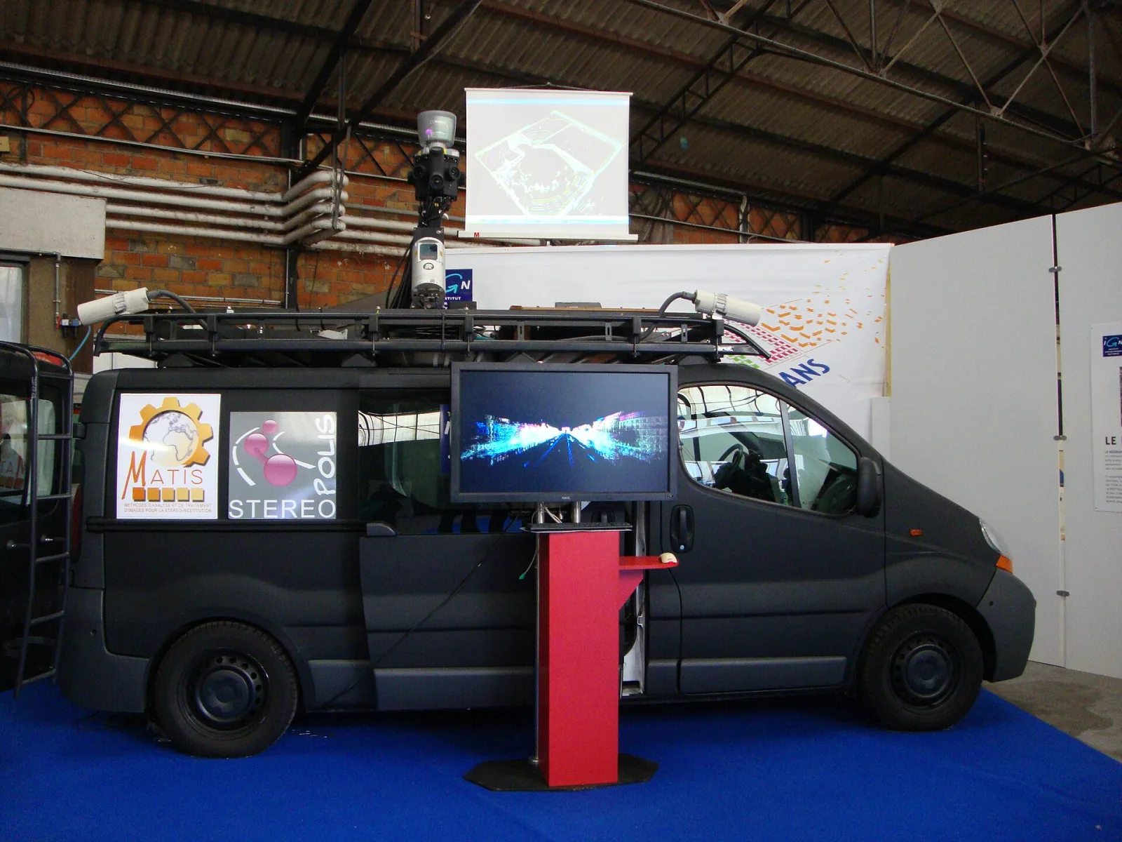 A dark van with a mast of cameras and a laser scanner on its roof, displayed at an exhibition, with the logos of the French IGN’s MATIS laboratory and Stéréopolis on its side
