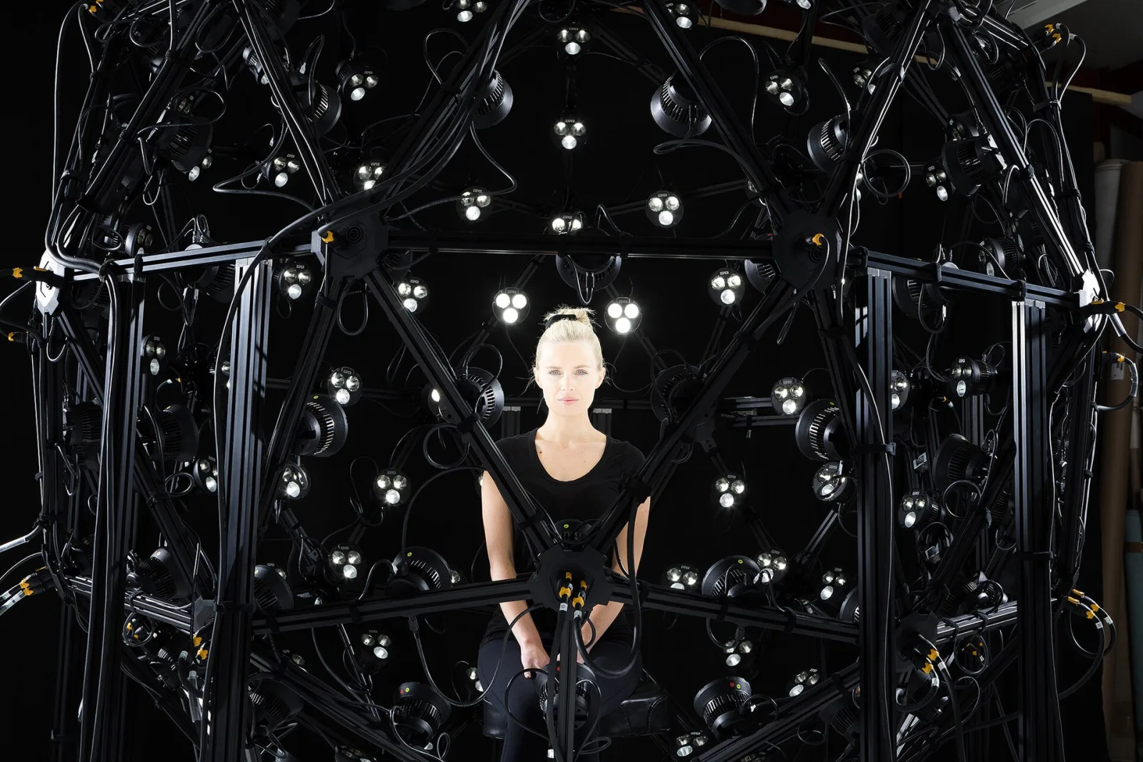 A photogrammetry cage: a dome of black metal tubes carrying dozens of cameras and lights, all pointing at the person seated at its centre